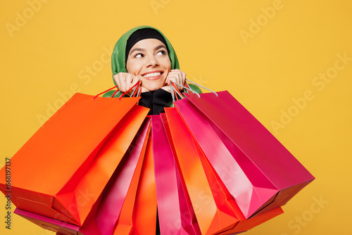 Young smiling cheerful arab asian muslim woman wears green hijab abaya clothes hold shopping package bags look aside isolated on plain yellow background . Black Friday sale buy day, uae islam concept. photo