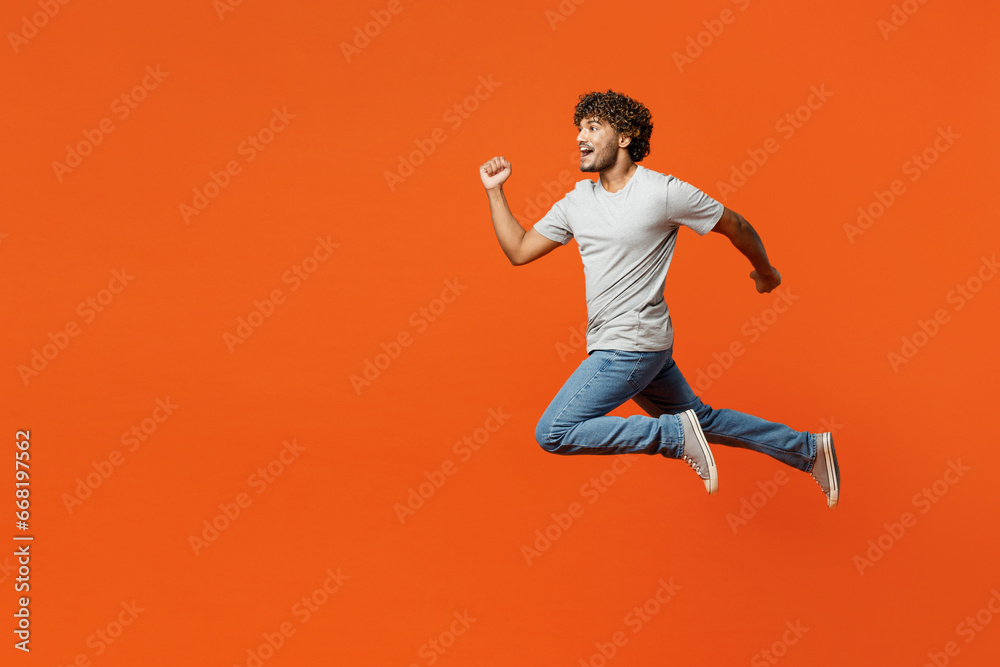Full body side view young overjoyed excited cool smiling happy Indian man wears t-shirt casual clothes jump high run fast isolated on orange red color background studio portrait. Lifestyle concept.