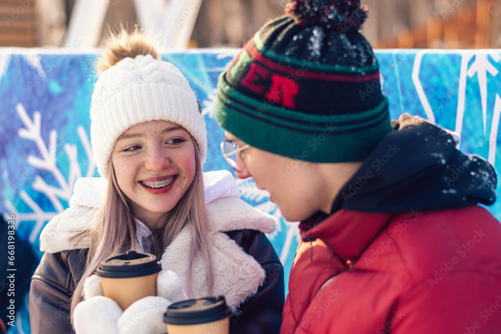 Obraz premium Young couple in love have fun, active date ice skating on the ice arena on a winter day.Laughing teenagers relax on a bench,drink hot cocoa,talk.Winter entertainment,leisure activity,Valentine's day.