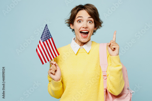 Young shocked woman student wears casual clothes yellow sweater backpack bag hold in hand American flag point index finger up isolated on plain blue background. High school university college concept.