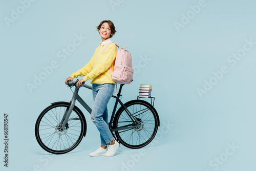 Full body young smiling happy cheerful woman student wear casual clothes sweater backpack bag ride bicycle look aside on area isolated on plain blue background. High school university college concept.