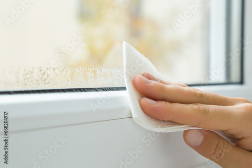 Young adult woman hand fingers holding dry white paper napkin and wiping water drops from glass and window plastic frame. Closeup. Room humidity problem. photo
