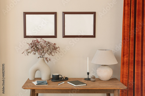 Two empty picture frames on a wall by a table with assorted ornaments, stationary, lighting and a vase filled with flowers photo