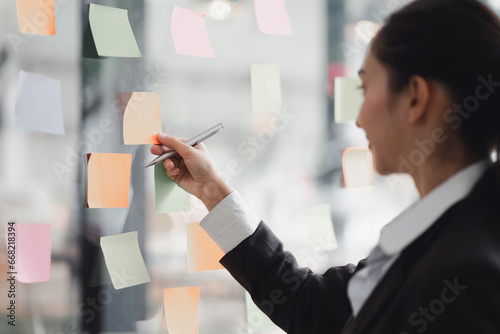 Businesswoman writing notes about teamwork decisions for project plan, tasks, or ideas on sticky notes in startup workplace.