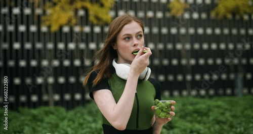 Young girl in sportswear with headphones is eating a fruit salad in the park.
