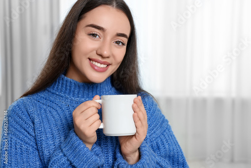 Happy young woman holding white ceramic mug at home, space for text