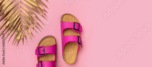 Fashionable leather bright pink birkenstock sandals, golden palm leaf on pink background, top view flat lay. Minimalistic concept of summer unisex shoes, flip-flop sandals for vacation, beach