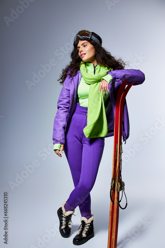 winter activity, charming woman with curly hair posing in active wear with puffer jacket and skis