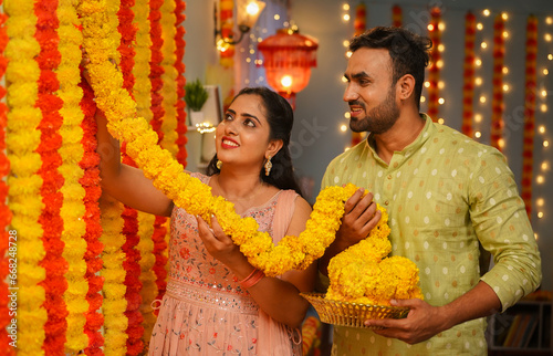 happy husband helping wife for doing flower decoration for diwali festival celebration at home - concept of festive preparation, family bonding, traditional culture or custom. photo