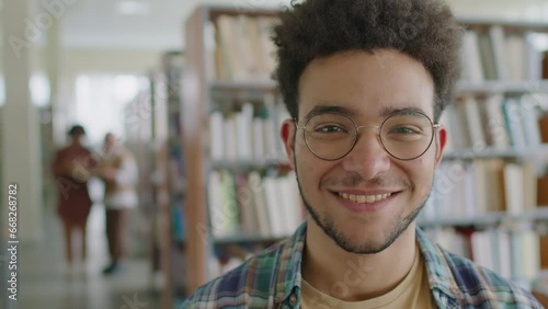 Medium closeup portrait of young Biracial male migrant student smiling at camera on blurred library background