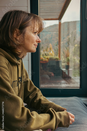 Girl sitting on a bed behind big window with autumn mountains view. Woman in hoodie on window sill. Escape in mountain cabin. Leisure in countryside cottage. Autumn tourism. Happy person in mountains.