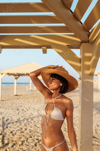 Smiling young woman wearing a hat enjoying her holiday on the beach. Satisfied beautiful girl relaxes on the beach during sunrise.