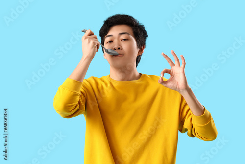 Young Asian man with spoon showing OK on blue background