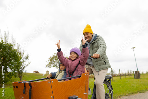 Mature man in warm casual clothes  drives a cargo bicycle  with two excited  little children on board in a contryside photo