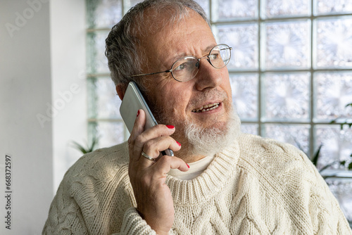 Mature man LGBTQ with grey hairs and colored nails  talking on cell phone at home against the glass wall 