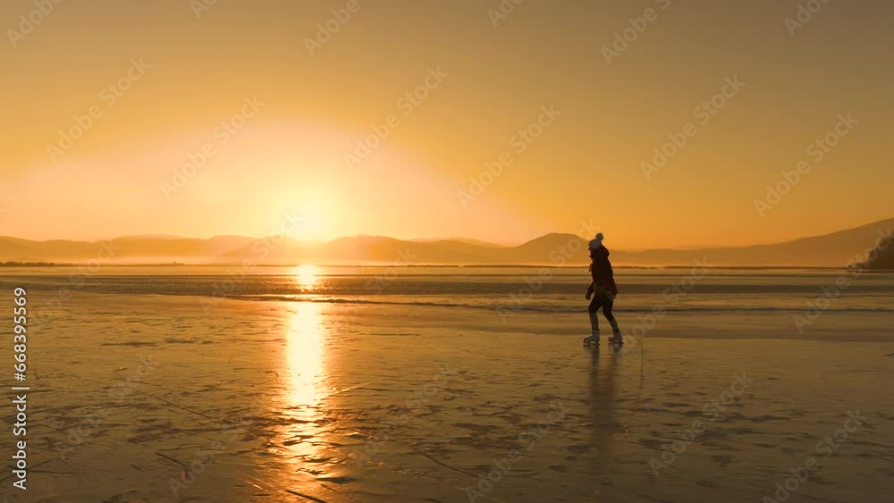 LENS FLARE, SILHOUETTE: Female ice skater gliding on golden glowing ice surface of a naturally frozen lake. Last rays of sun reflect from ice while she skates across big pond surrounded with hills. LENS FLARE, SILHOUETTE: Female ice skater gliding on golden glowing ice surface of a naturally frozen lake. Last rays of sun reflect from ice while she skates across big pond surrounded with hills.