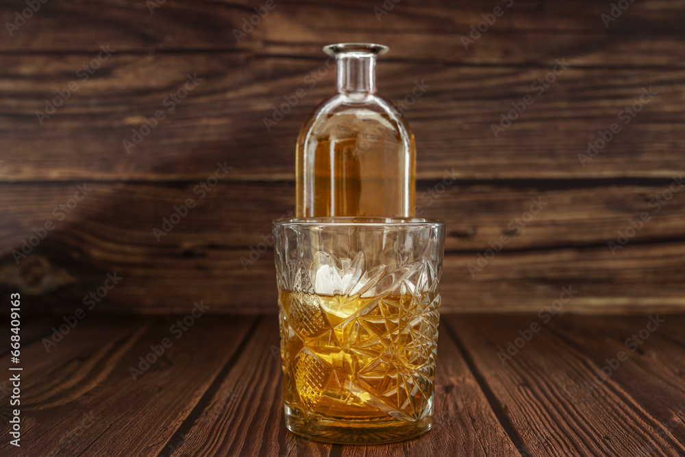A bottle with liquor next to a carved glass filled with liquor and ice on a dark wooden surface A bottle with liquor next to a carved glass filled with liquor and ice on a dark wooden surface