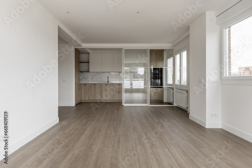 Empty living room with large windows, synthetic wood flooring and kitchen in the back with retractable screen