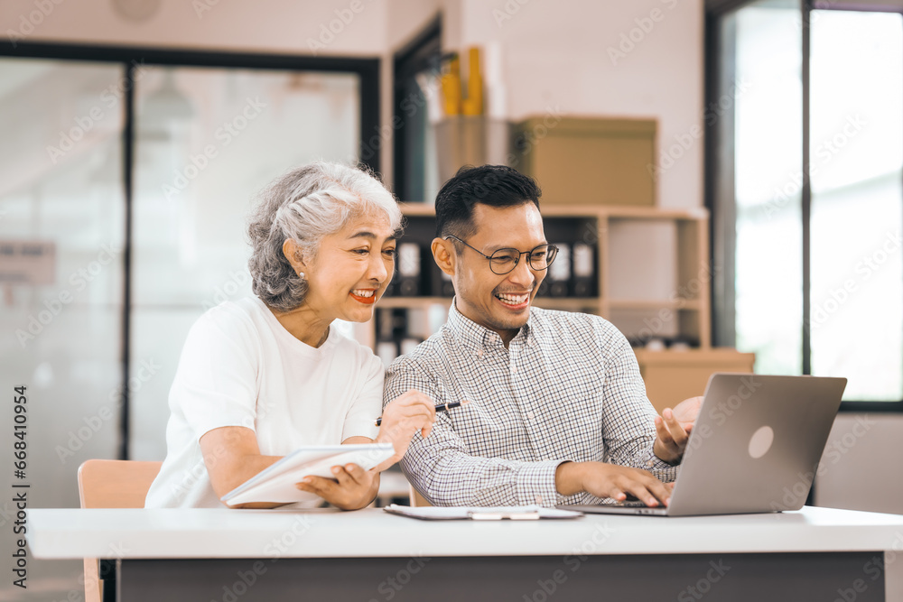 Fototapeta premium An older mature woman shows something to asian man on a laptop. They smile and work together in an office. They look happy and focused.