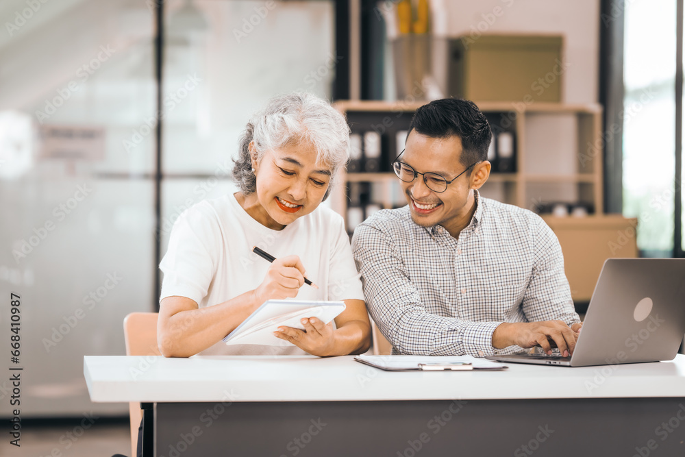 An older mature woman shows something to asian man on a laptop. They smile and work together in an office. They look happy and focused. An older mature woman shows something to asian man on a laptop. They smile and work together in an office. They look happy and focused.