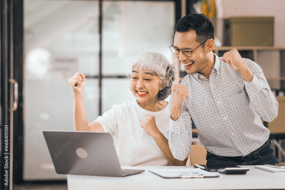 An older mature woman shows something to asian man on a laptop. They smile and work together in an office. They look happy and focused. An older mature woman shows something to asian man on a laptop. They smile and work together in an office. They look happy and focused.