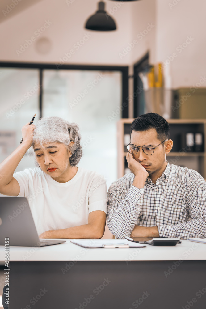 An older mature woman talks to asian man. He covers his face with his hands. They are in an office. They have a laptop and papers. The man looks stressed. An older mature woman talks to asian man. He covers his face with his hands. They are in an office. They have a laptop and papers. The man looks stressed.