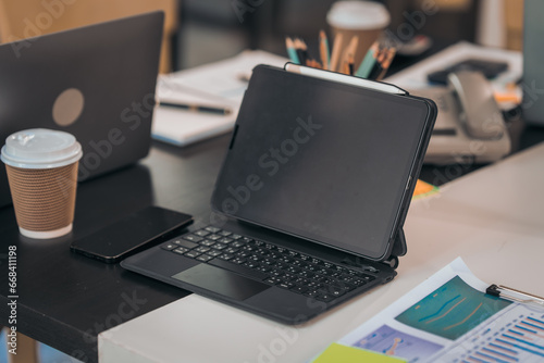 A laptop on a desk displays charts. There's a calculator, coffee, papers, and pens. The workspace seems organized and busy.