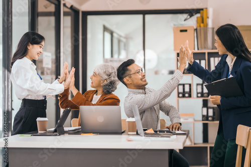 Four people in an office are happy and celebrating. They give high-fives and clap hands. They sit and stand near a table with laptops and coffee. discuss opinions, presentation teamwork
