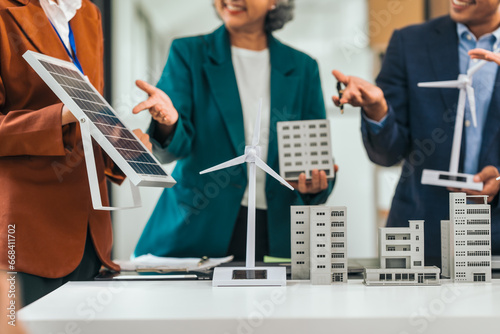 Four asian businesspeople professionals discuss renewable energy in office, showing solar panel and wind turbines with tower building model, apartment. modern business with environmental, clean energy