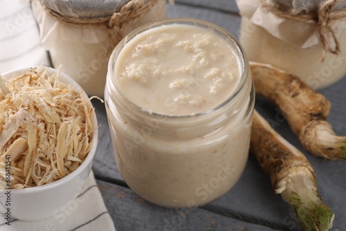 Spicy horseradish sauce in jars and roots on grey wooden table, closeup
