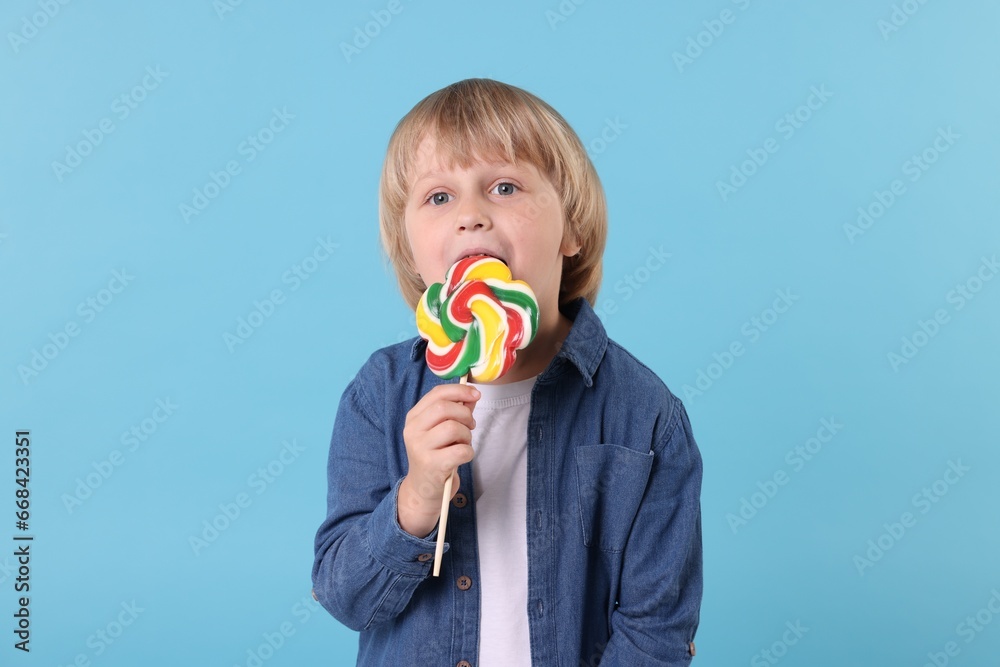 Cute little boy licking colorful lollipop swirl on light blue background Cute little boy licking colorful lollipop swirl on light blue background
