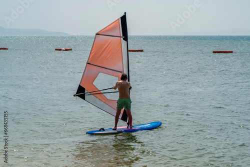 A man in green shorts rides a blue board with a yellow sail in sunny weather at a tropical resort. He stands on the board, holding onto the sail.
