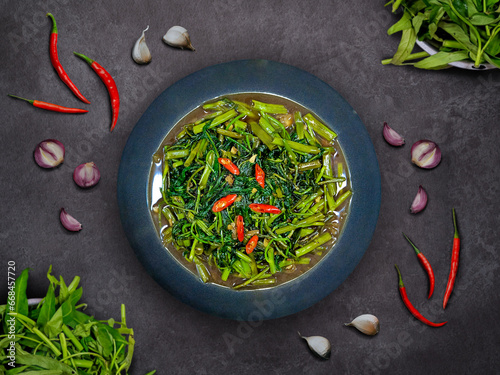Stir fried water morning glory on a dark plate with garlics, chilis, and shallots scattered around the plate. Known as oseng or tumis kangkung to Indonesian. photo