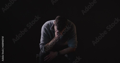 Sitting, sad man sobs in dark room, black background. Static