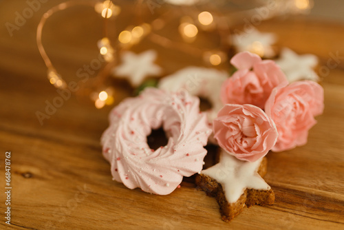 Gingerbread cookies in the shape of stars, merengue cookies and pink roses on a wooden table against a background of bokeh of New Year’s lights. Christmas background. Winter card. Delicious pastries © Liudmila
