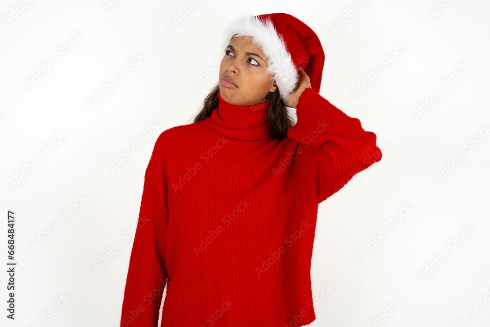 Beautiful girl wearing red sweater and Santa hat confuse and wonder about question. Uncertain with doubt, thinking with hand on head. Pensive concept. Beautiful girl wearing red sweater and Santa hat confuse and wonder about question. Uncertain with doubt, thinking with hand on head. Pensive concept.