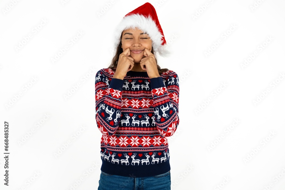 Pleased Beautiful girl wearing christmas sweater and Santa hat with closed eyes keeps hands near cheeks and smiles tenderly imagines something very pleasant Pleased Beautiful girl wearing christmas sweater and Santa hat with closed eyes keeps hands near cheeks and smiles tenderly imagines something very pleasant