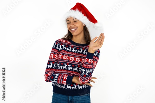 Overjoyed successful Beautiful girl wearing christmas sweater and Santa hat raises palm and closes eyes in joy being entertained by friends