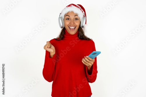 Positive Beautiful woman wearing christmas hat holds modern cell phone connected to headphones, clenches fist from good emotions, exclaims with joy,