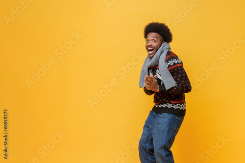 Happy young African-American man in Christmas sweater and scarf expressing surprise feeling in isolated yellow background with copy space studio shot photo