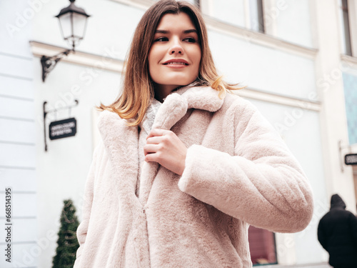 Young beautiful smiling lady wearing trendy white faux fur coat. Stylish woman posing in the street in winter. Cheerful and happy model in. Cold autumn weather. Shopping mall