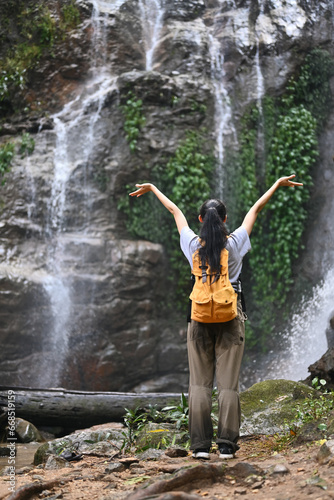 Excited female traveler with backpack raising arms relaxing enjoying at tropical nature waterfall