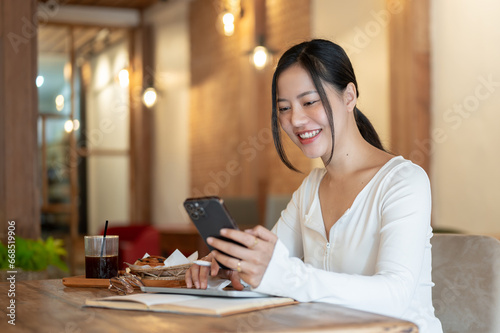 A happy Asian woman is using her smartphone, texting her friends while sitting in a coffee shop.