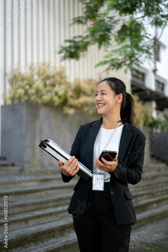 A businesswoman stands outside of the building with a smartphone and document binders in her hands.