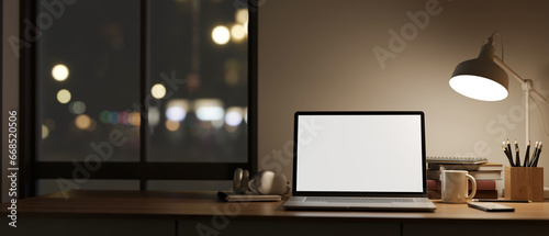 Fototapeta Naklejka Na Ścianę i Meble -  A white-screen laptop mockup on a table against the window and wall in a modern dark room at night.