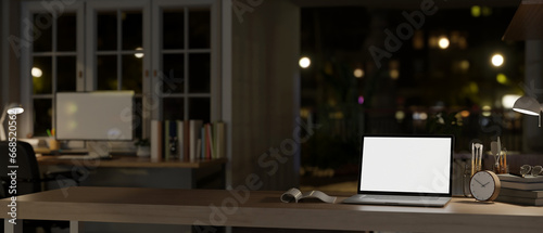 A modern dark office at night with a white-screen laptop mockup and accessories on a tabletop.