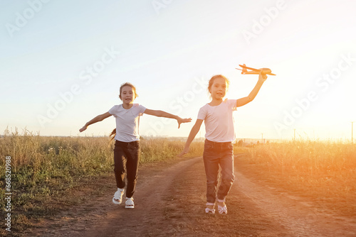 Joyful sisters run along road in field playing with toy plane. Concept of entertainment in childhood and imitation of flight. Imagination and play
