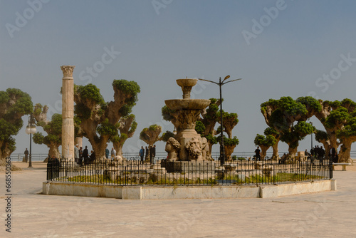 Cherchell, Algeria, 10 23 2023 : main square of Cherchell in a misty atmosphere. Algerian people, sand, plane trees, Roman column, fountain. photo