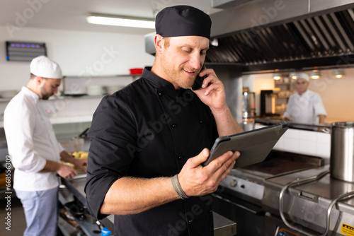 Caucasian male chef teacher using tablet and talking over cellphone with students in background photo
