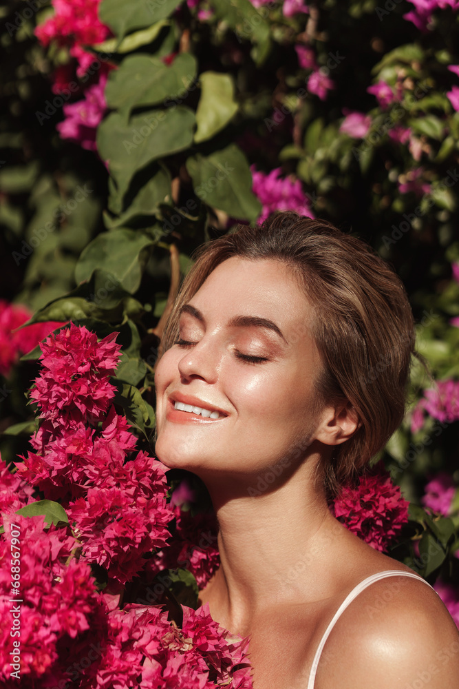 Fototapeta premium Beautiful blonde woman with smooth skin and natural makeup, as she poses against a backdrop of a bush with delicate pink flowers